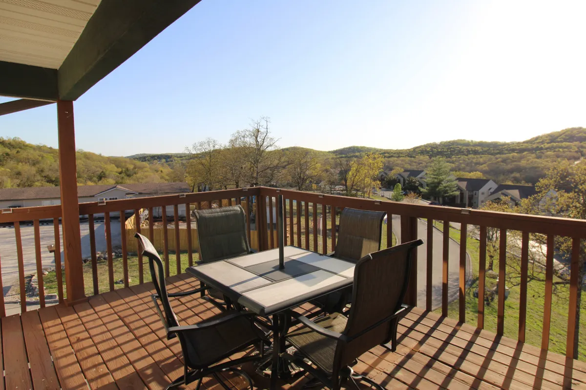 This inviting exterior features a table and chairs set on a wooden deck, overlooking expansive hills and greenery. The space is perfect for outdoor dining, morning coffee, or evening gatherings with family, set against a backdrop of natural beauty.