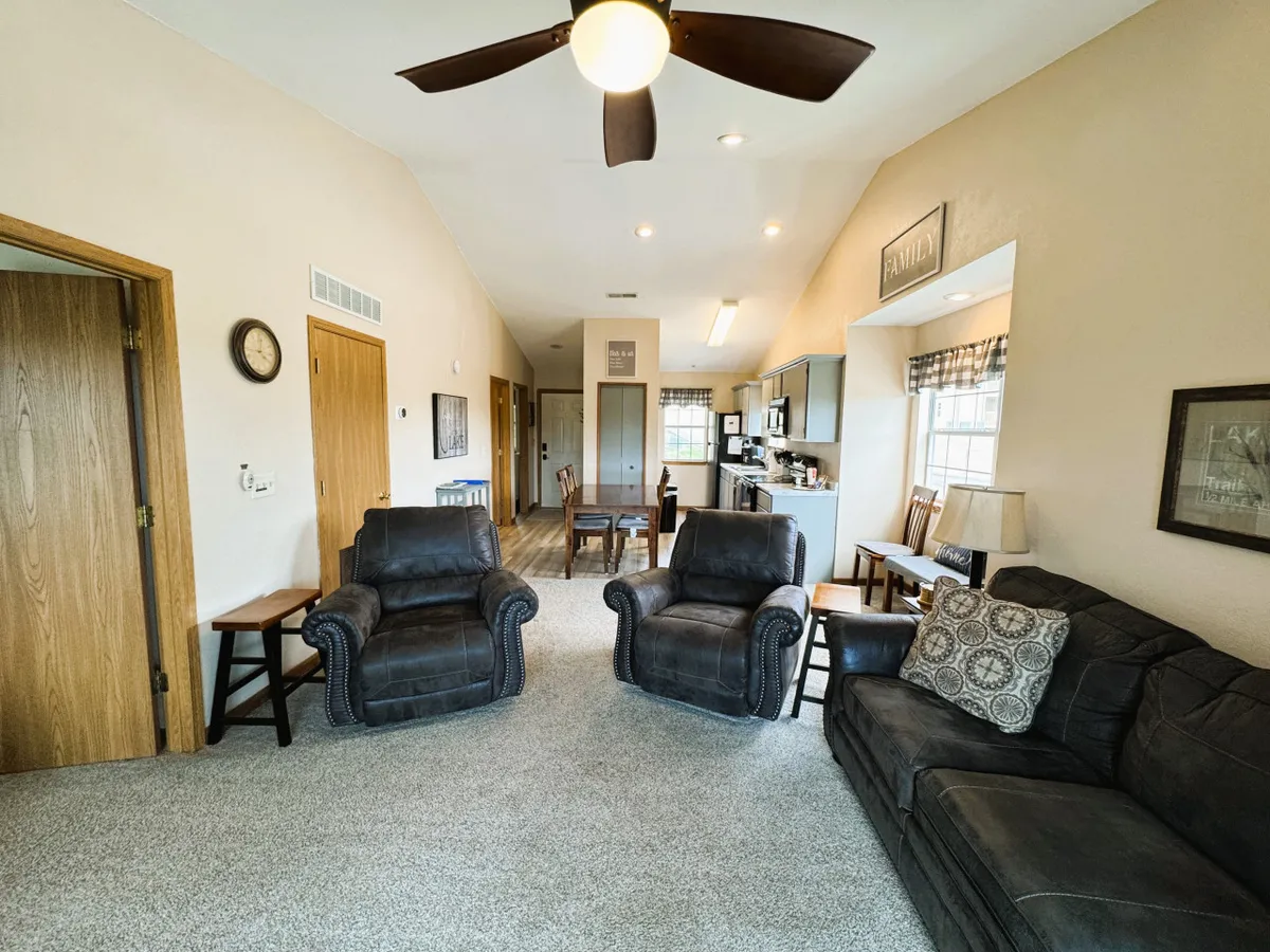 This image showcases an inviting open-plan living space with comfortable armchairs, a plush sofa, a dining table, and a clear view into the adjacent kitchen area. Decorative wall art and warm lighting further enhance the welcoming, homey feel of the room.