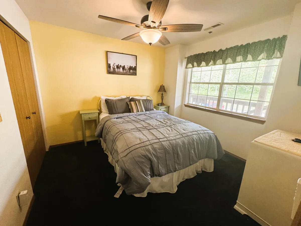 A cozy bedroom with a neatly made bed, large window letting in morning light, a ceiling fan for comfort, a bedside lamp, and decorative wall art. The space feels inviting and restful, perfect for unwinding after a busy day.