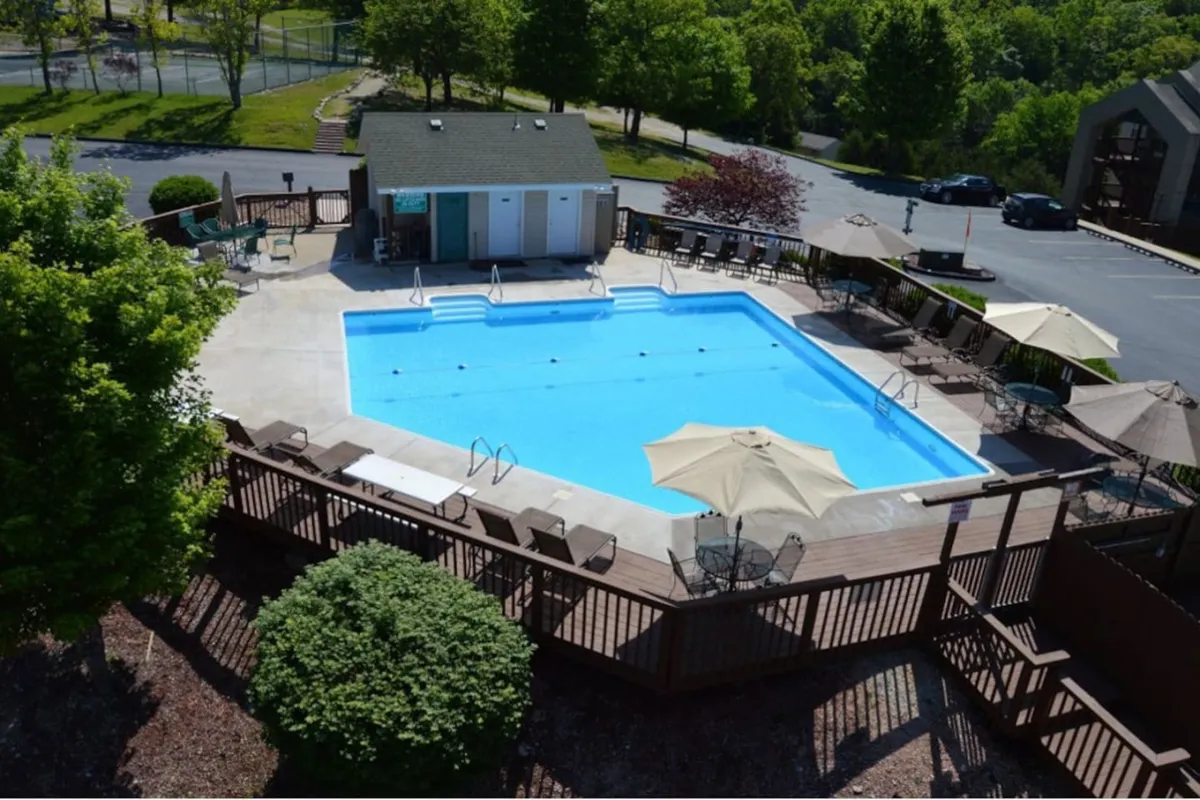 A vibrant pool area bordered by a wooden deck, with lounge chairs and umbrellas placed for ultimate relaxation. The sparkling water, greenery, and seating encourage sunbathing, swimming, and social interaction in a resort-like atmosphere.