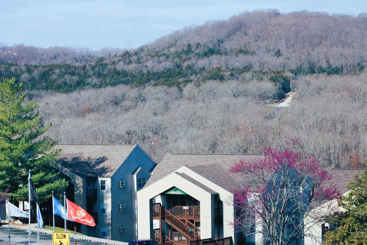 A tranquil exterior shot with mountains in the background, buildings nestled among trees, and flags marking a community area. The scene captures guests engaging in hiking, relaxing, and enjoying community events, with sunset light enhancing the inviting mood.