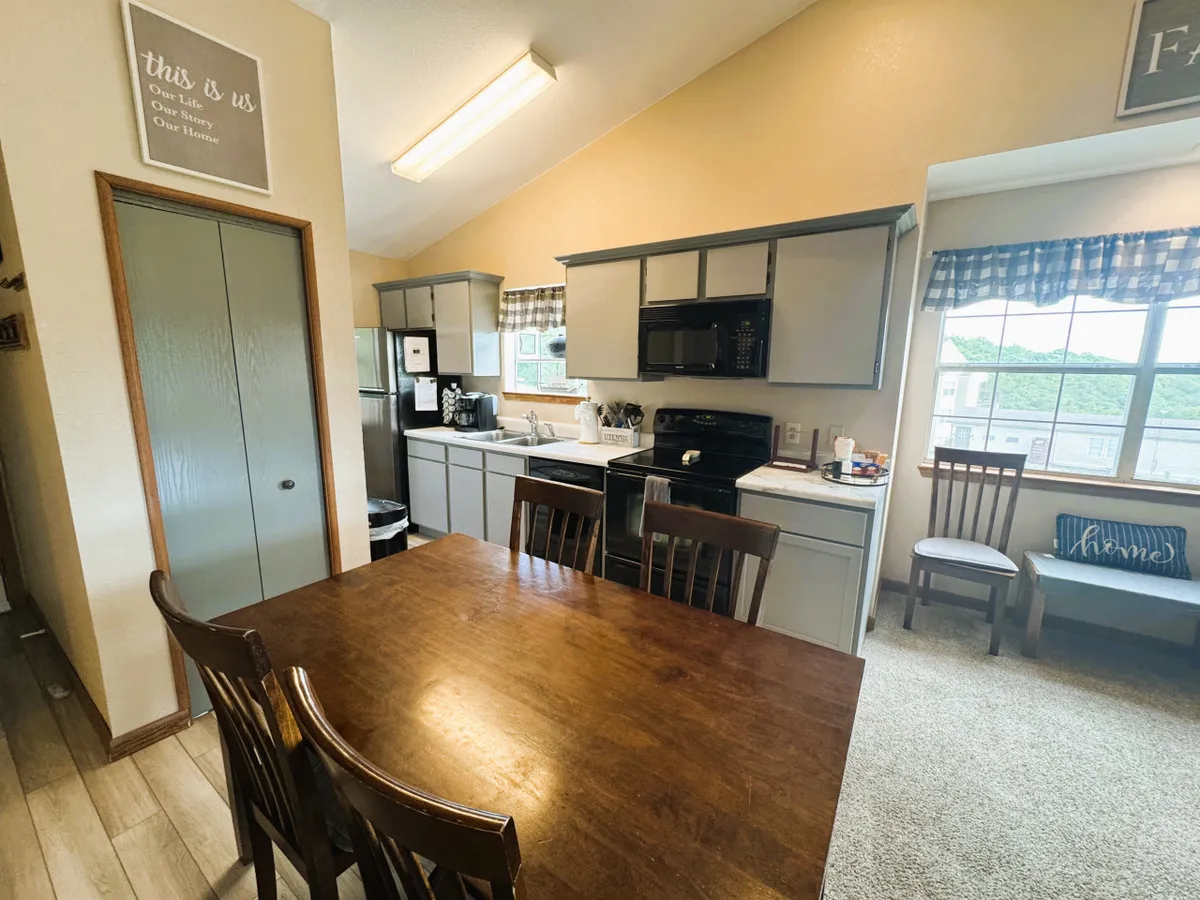 A warm and welcoming interior scene featuring a spacious wooden dining table, modern kitchen appliances, a large window letting in natural light, and a decorative sign. The image captures both the kitchen and dining area in one view, showcasing the ideal setting for family meals, cooking, and social gatherings.