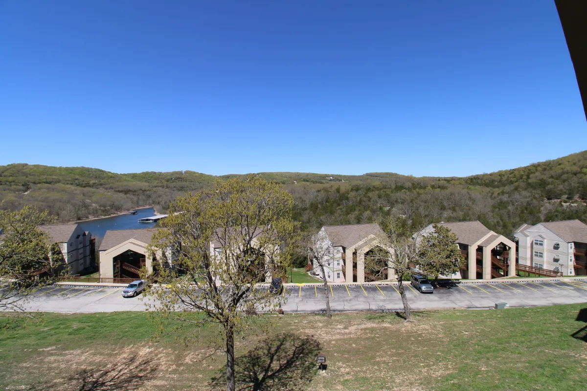 A peaceful lakeside setting with apartment buildings nestled among trees and gently rolling hills. The scene includes the sparkling lake and a serene natural environment, ideal for relaxing, boating, or enjoying the sunset.